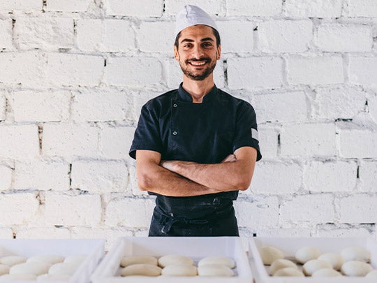 Emprendedor gastronómico sonriendo con los brazos cruzados frente a cestas de masas para panadería