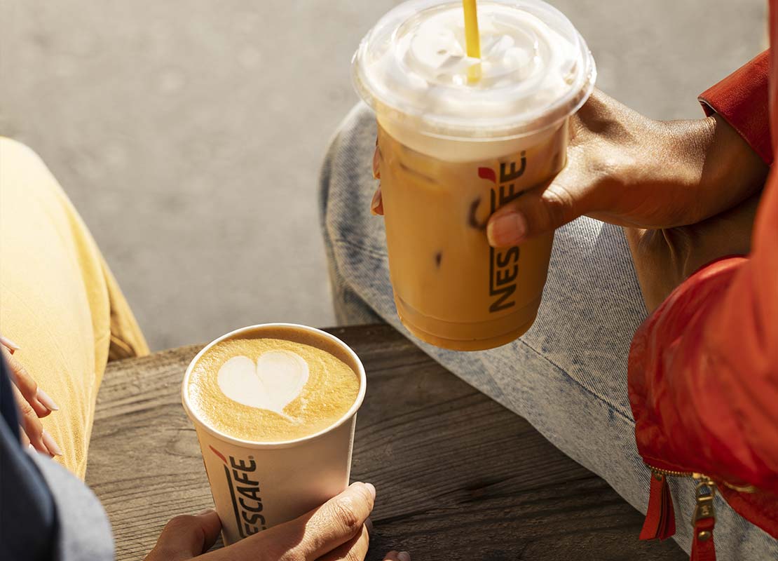 Sobre una tabla, una mano sostiene un vaso reciclable de NESCAFÉ® y otra un vaso plástico con café frío