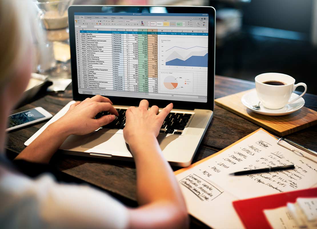 Manos de mujer teclean en una laptop que proyecta tablas y gráficos, al lado, planillas y facturas