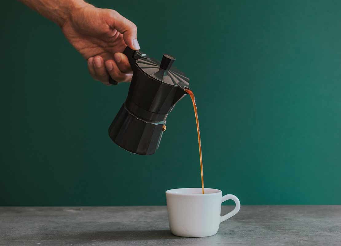 Mano sostiene una cafetera que vierte a una taza café preparado por método de percolación
