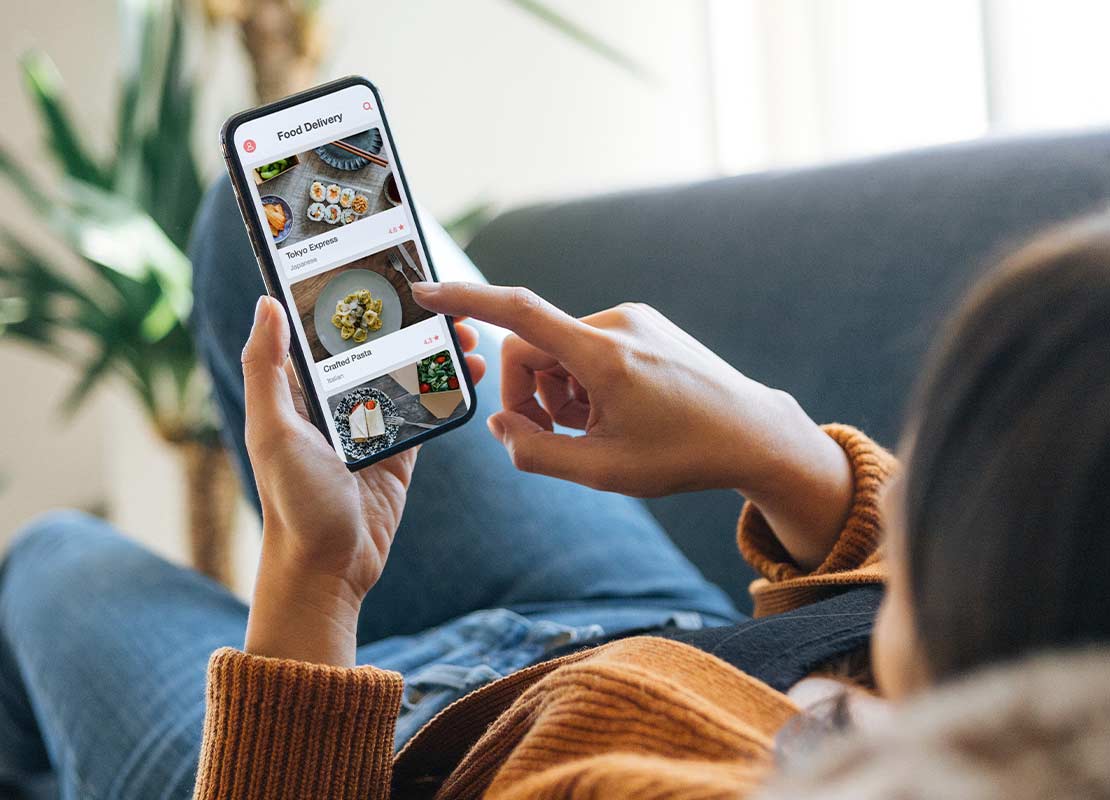 Mujer recostada en un sillón revisa una plataforma de entrega de comida a domicilio desde su celular