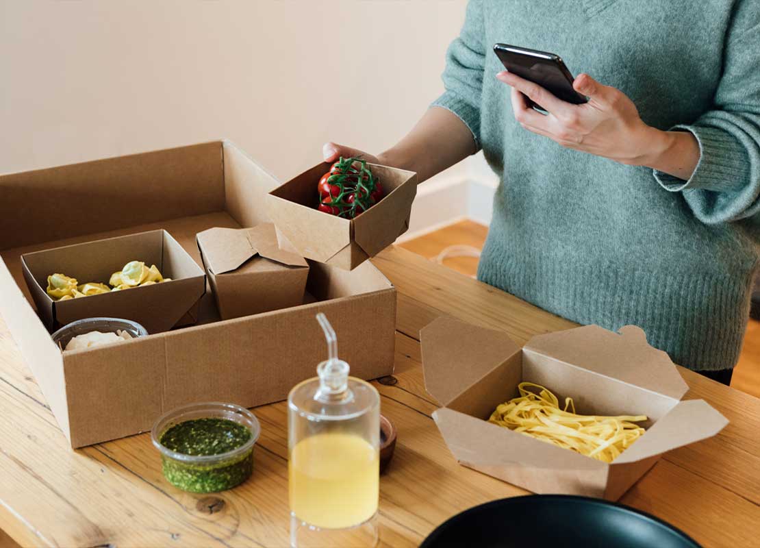 Hombre sostiene caja de comida en su mano y en la otra su celular, frente a él una mesa con un delivery
