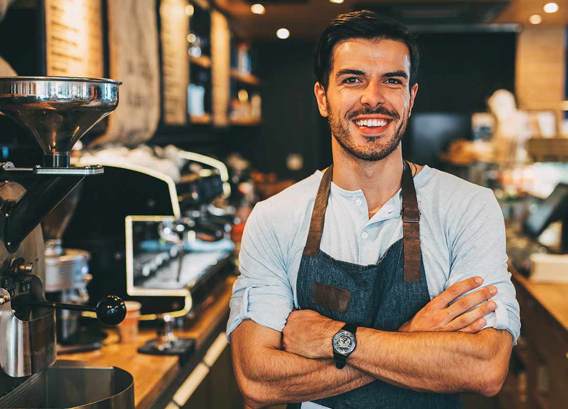 Barista cruzado de brazos sonríe. Al fondo sus máquinas y herramientas para elaborar café