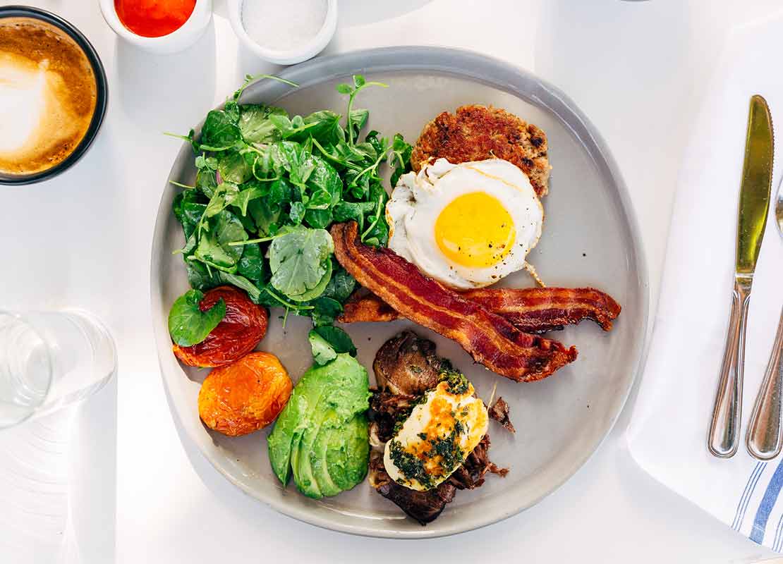 Plato con huevos, tocino, espinaca y carne, acompañado de cappuccino y salsas para un desayuno