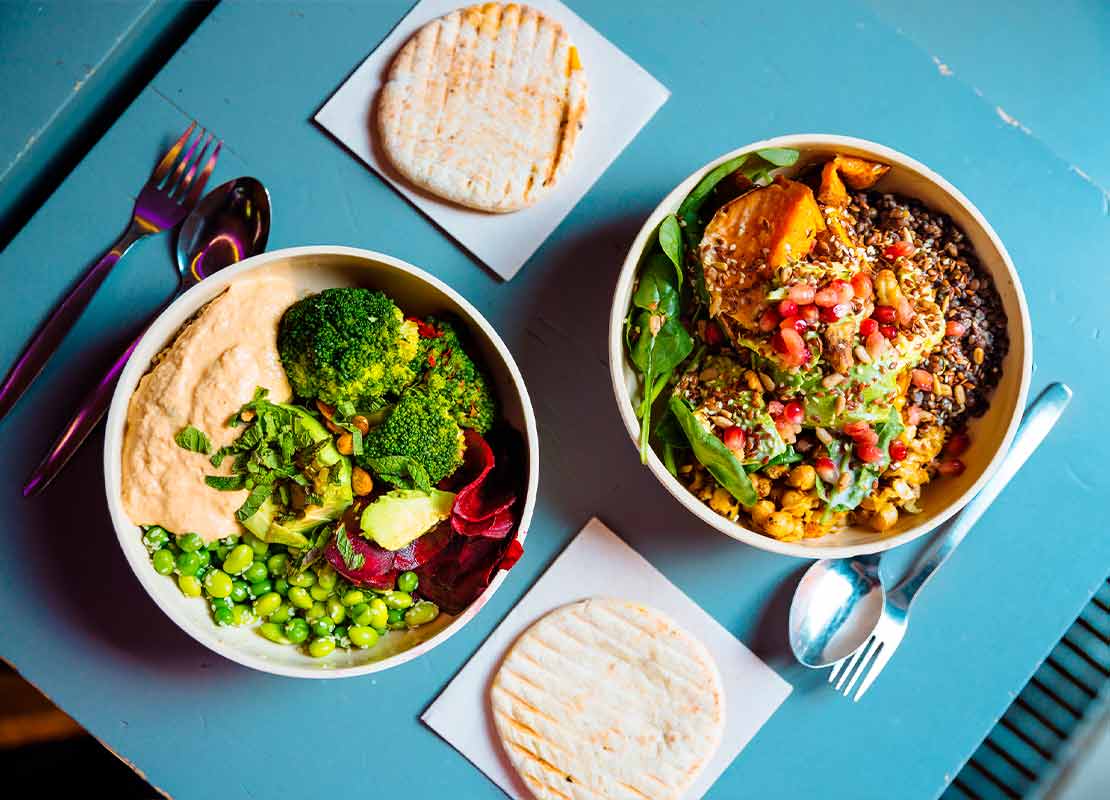 Dos bowls veganos con diversas verduras, granos y semillas, al lado, cubiertos y arepas