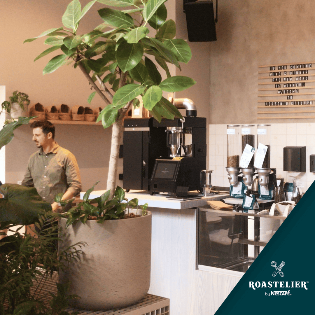 Hombre de pie frente a tostadora de café ROASTELIER™ con granos de café y una planta decorativa a su lado