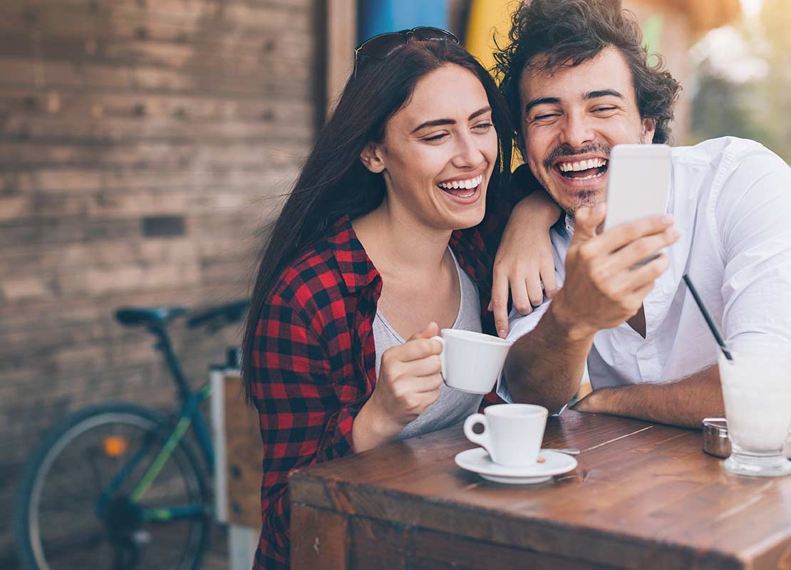 Pareja sentada a la mesa con tazas de café fuera de un restaurante sonríe mientras observa un smartphone