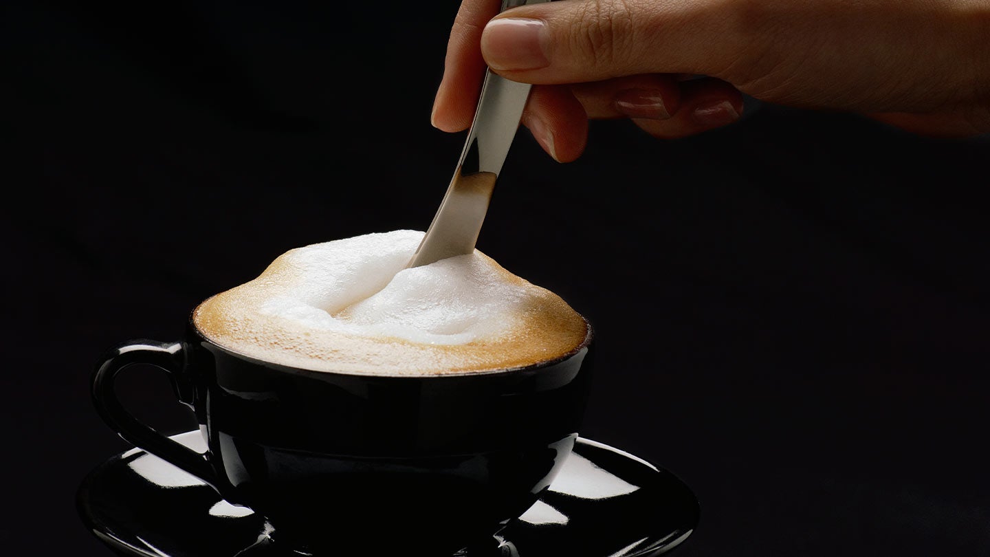 Mano de una mujer mezcla con una cuchara un cappuccino NESCAFÉ® servido en una taza negra
