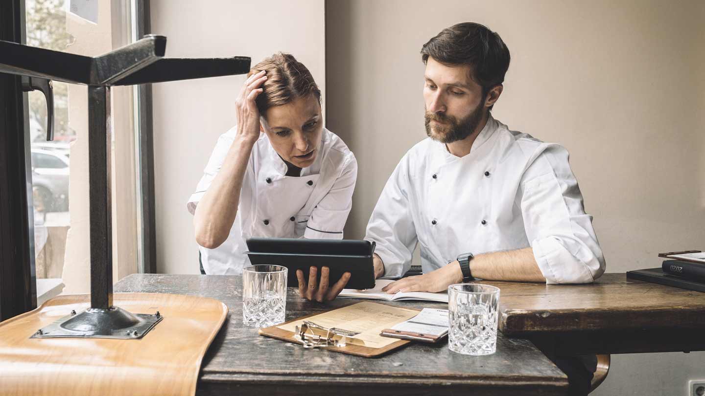 Dos chefs observan una tablet sentados a la mesa donde hay vasos, libretas y tabla legajadora