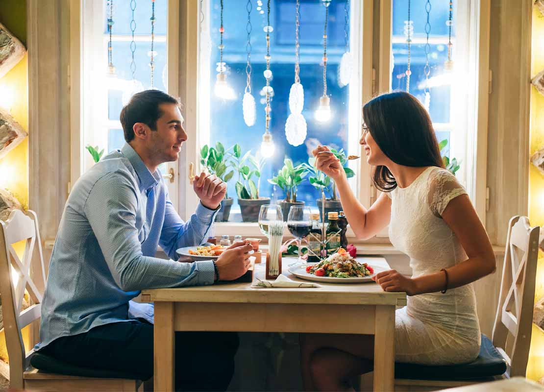 Pareja sentada a la mesa de un restaurante cena y conversa, al fondo una ventana iluminada con bombillos