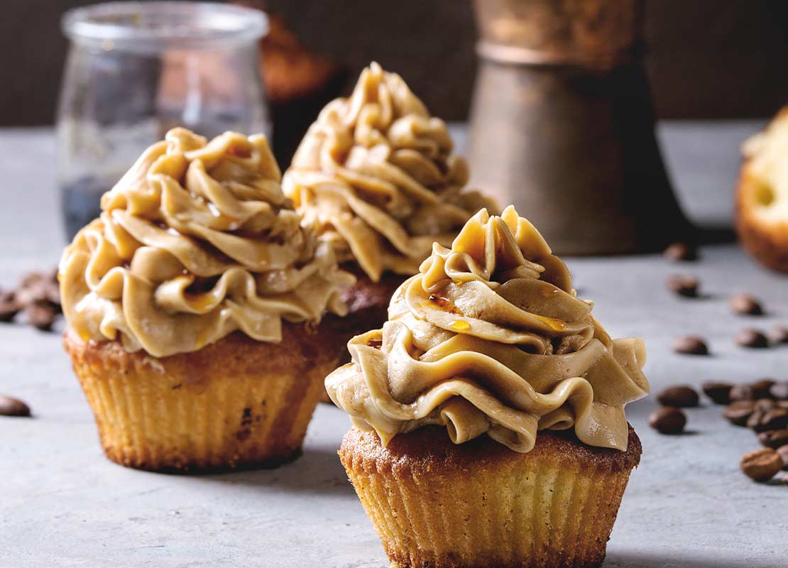 3 cupcakes decorados con crema de café y frutos secos, rodeados por granos de café NESCAFÉ®