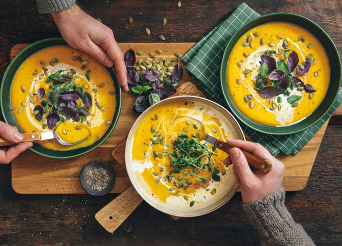 Manos mezclan con cucharas tres platos de sopas de vegetales servidas sobre una tabla de cortar