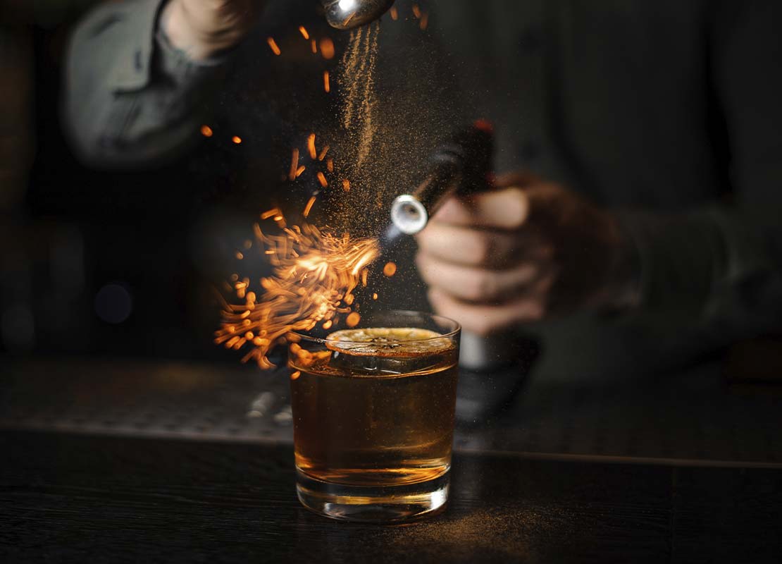 Manos de un mixólogo flamean canela en polvo que se vierte sobre una bebida en la barra de un negocio