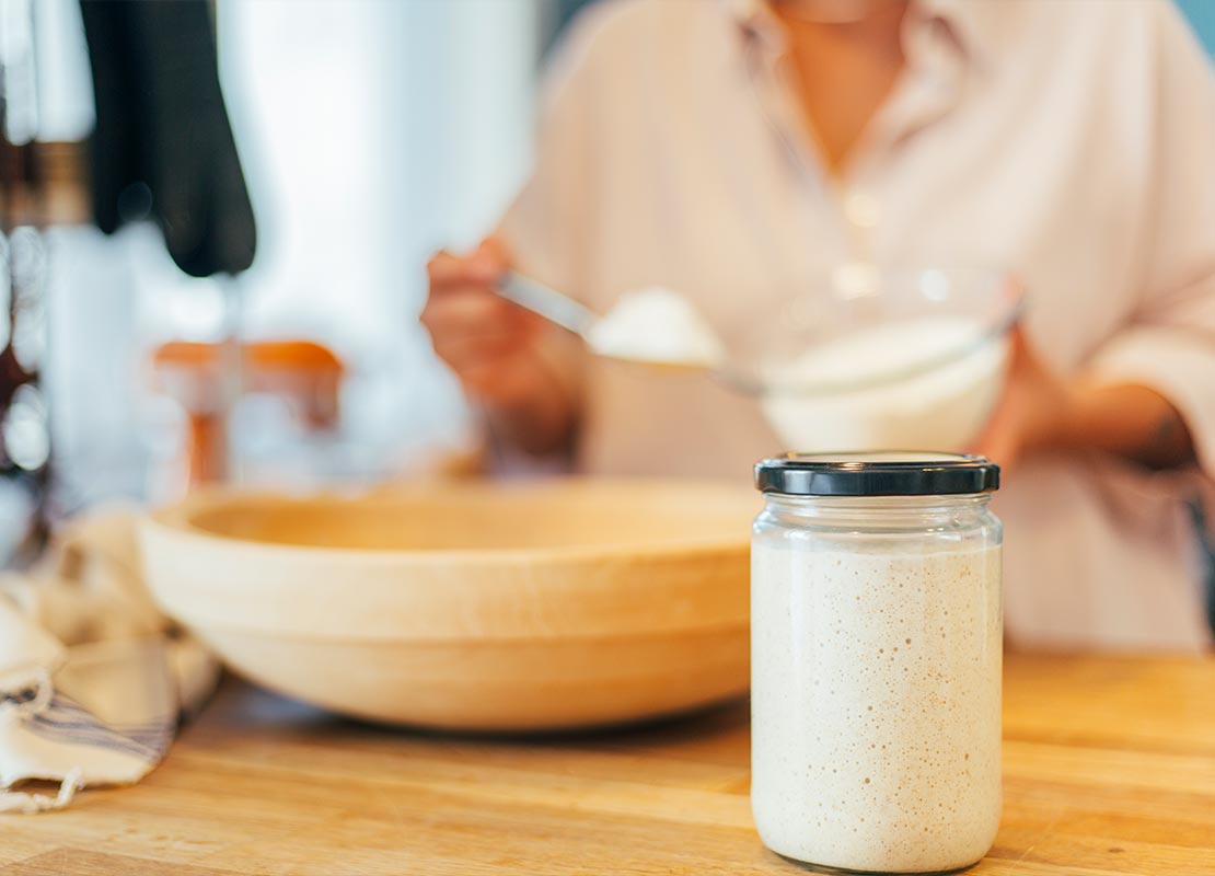 Frasco cerrado con levadura activa, al fondo mujer mezcla ingredientes en un tazón de madera