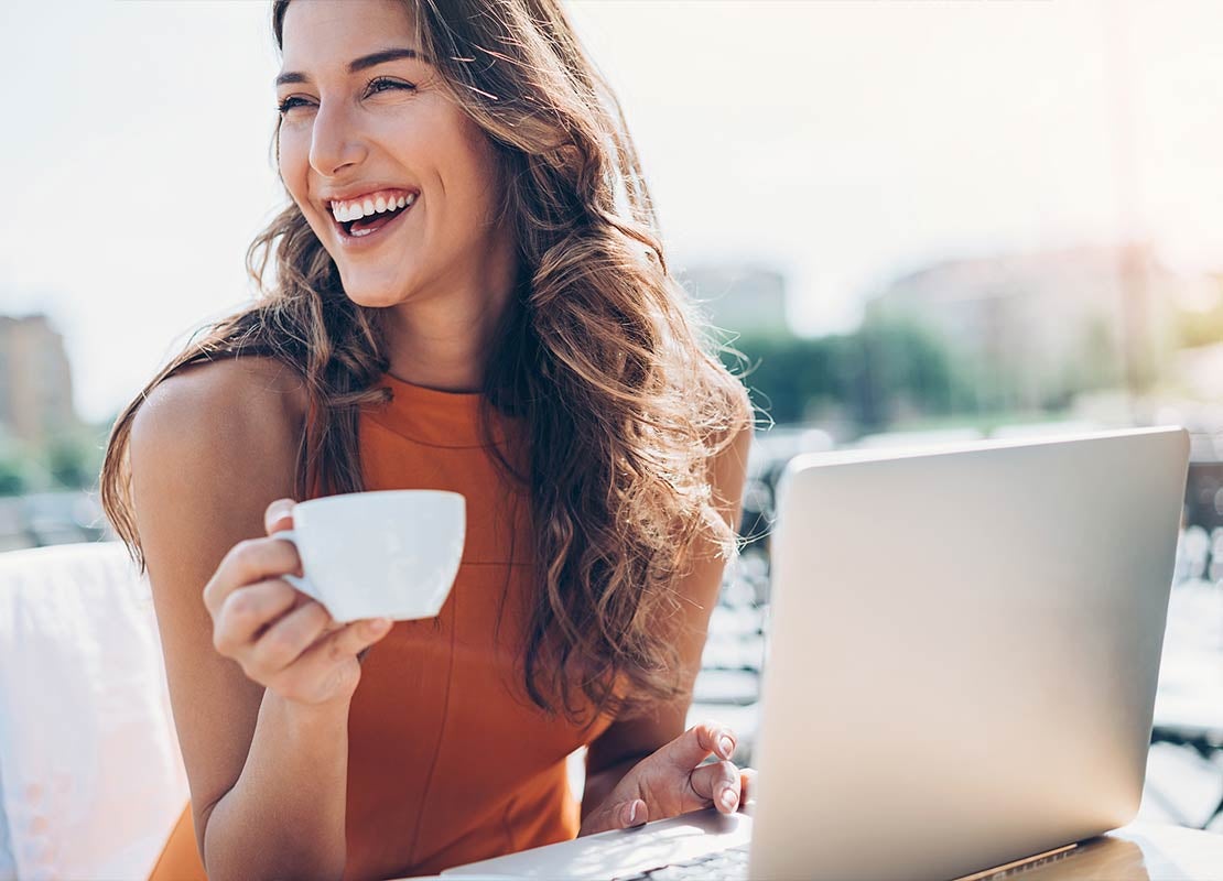 Mujer sonríe mientras sostiene una taza de café frente a un portátil de su trabajo al aire libre
