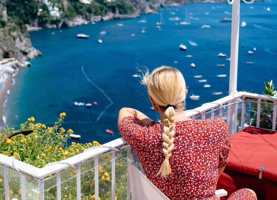 Mujer sentada a la mesa mira desde la terraza de un sky bar el mar, la playa y los botes a lo lejos