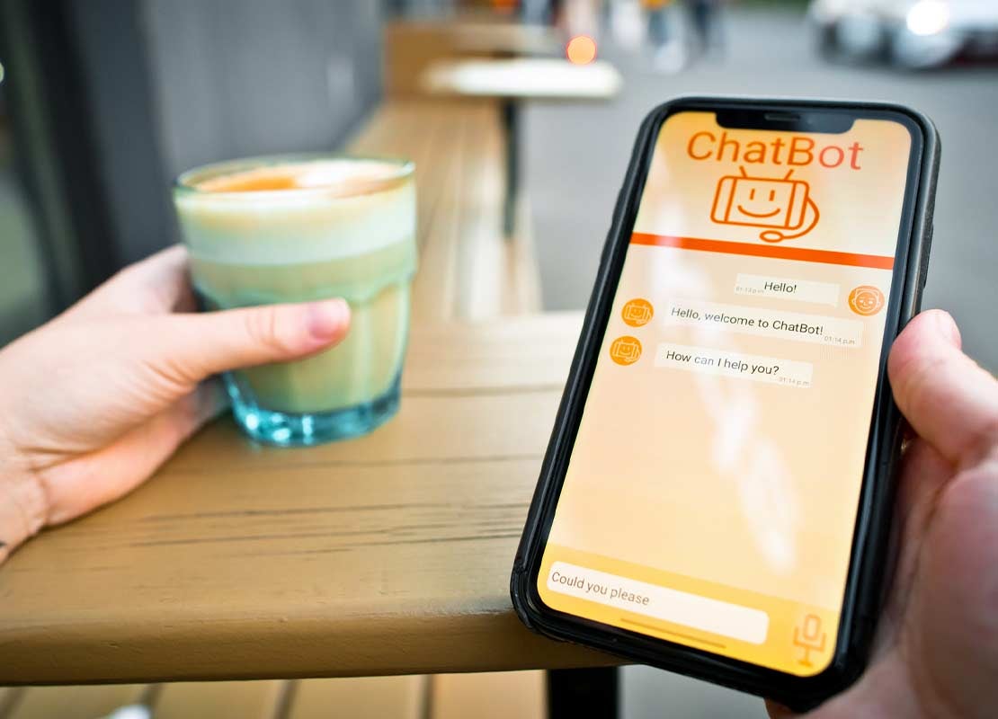 Mano sostiene un celular con un chatbot en su pantalla mientras con la otra mano sujeta un café
