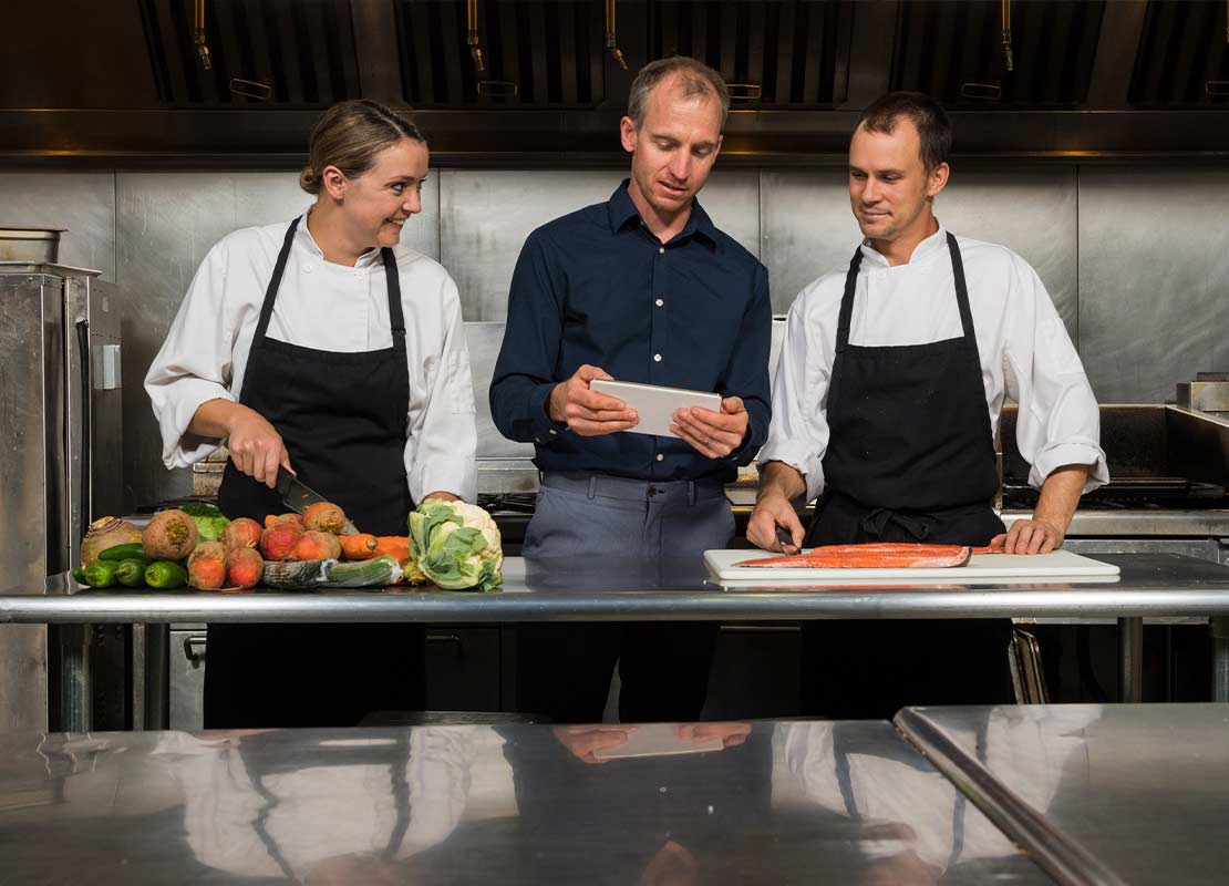 Dueño de restaurante y dos chefs observan una tablet, al frente una mesa con alimentos en una cocina comercial