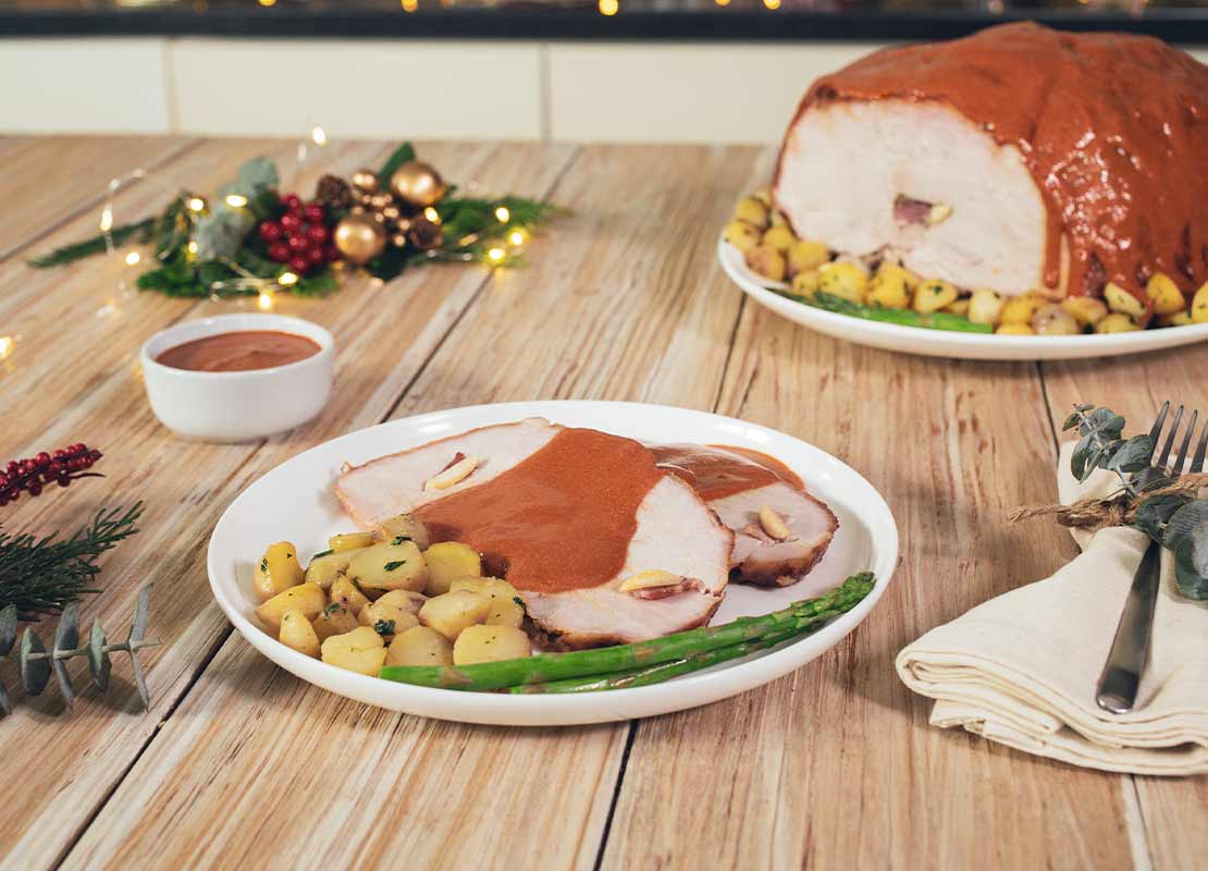Sobre una mesa, un plato con un trozo de pavo bañado en salsa, papas y verduras, al fondo detalles navideños