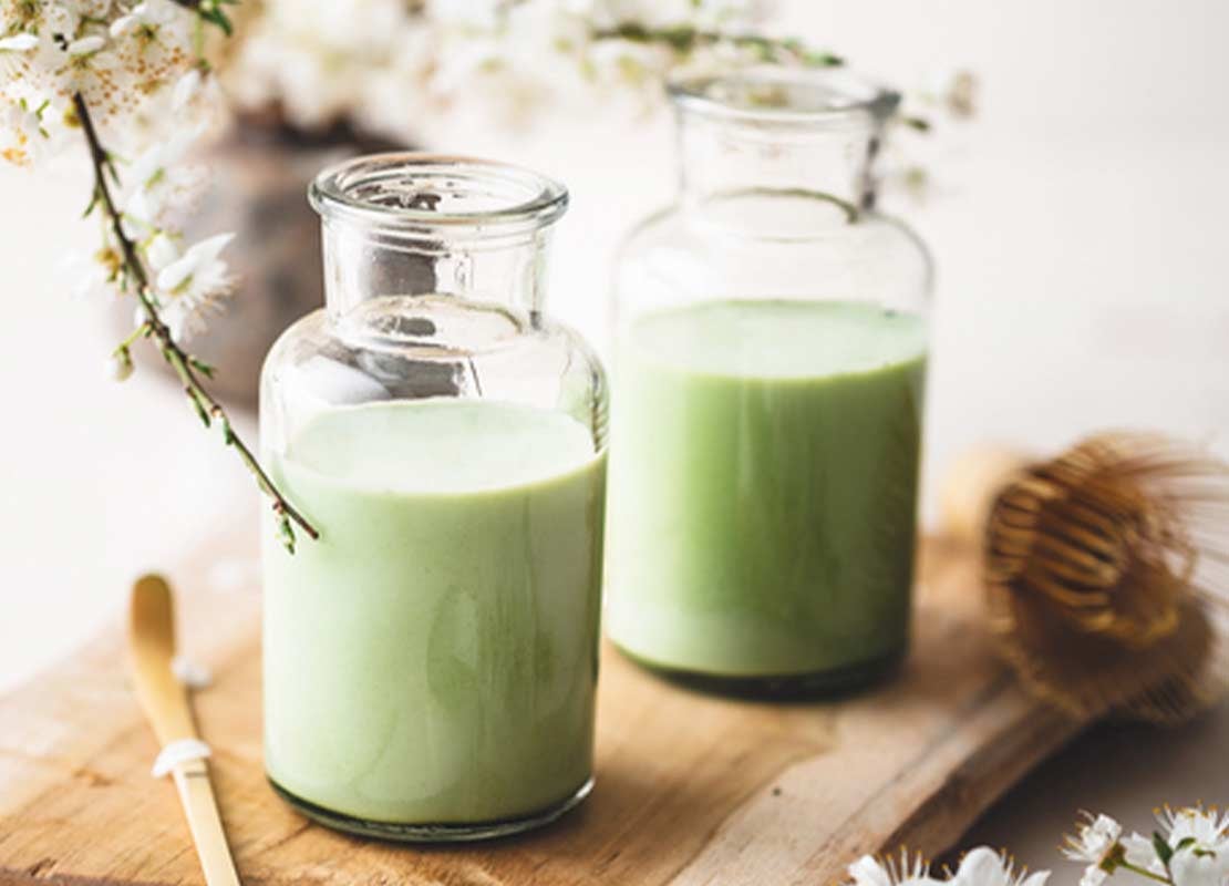 Sobre una tablan de cortar, dos botellas con Matcha Latte, al lado el ingrediente principal y flores