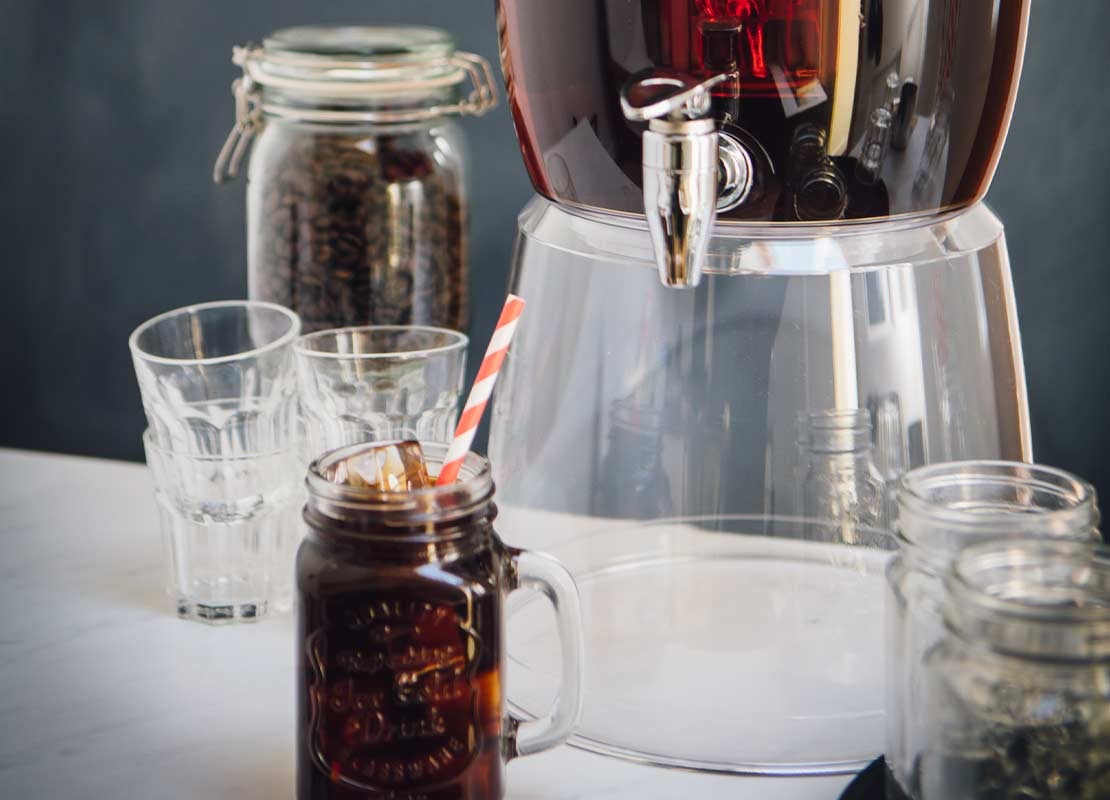 Dispensador y jarros de cristal con cold brew coffee y hielo, al lado frascos con granos de café