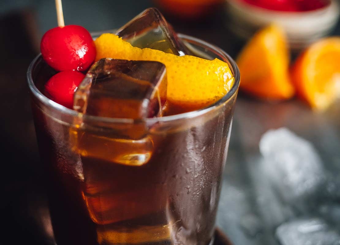 Vaso con una sangría sin alcohol, cubos de hielo, cerezas, un trozo de naranja, al fondo los ingredientes