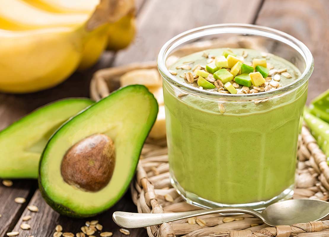 Vaso de bebida vegana de aguacate con avena en copos, al lado el fruto en mitades y plátanos