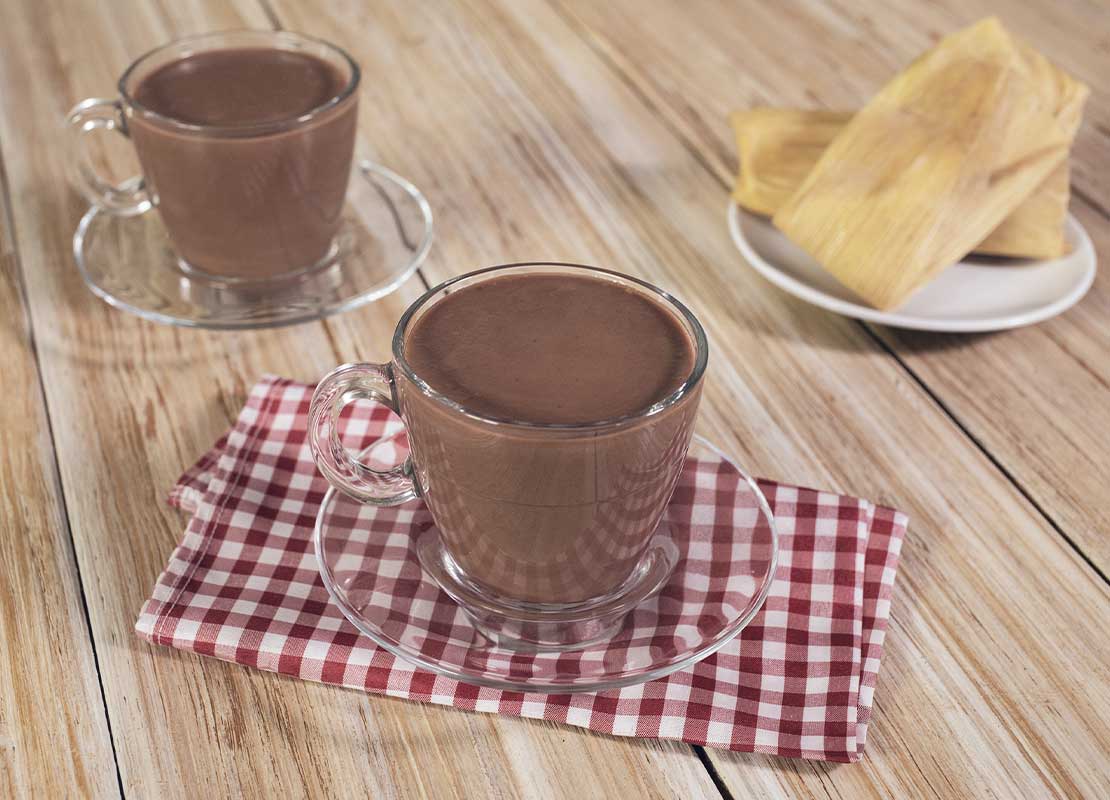 Dos tazas de cristal y sus platos con chocolate caliente, al lado, tamales de elote