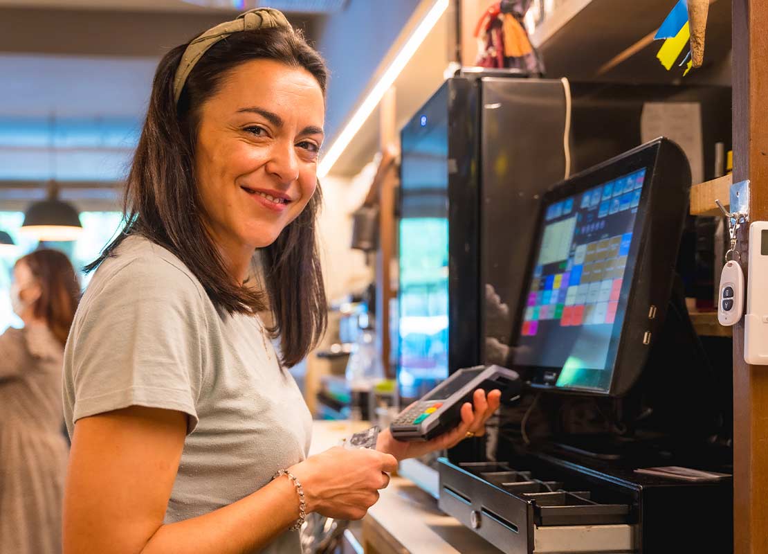 Cajera sonriente sostiene un datafono frente a computador y registradora de punto de venta de un negocio