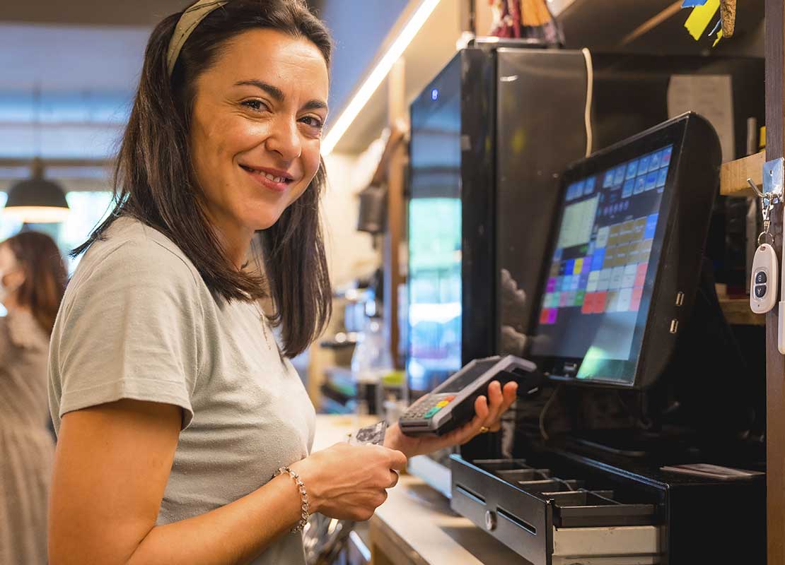 Cajera sonriente sostiene un datafono frente a computador y registradora de punto de venta de un negocio