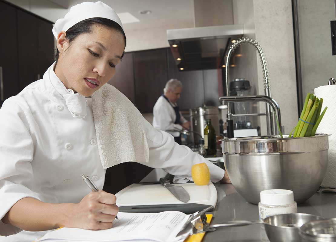 Chef escribe en una planilla sobre un mesón de cocina comercial mientras manipula utensilios