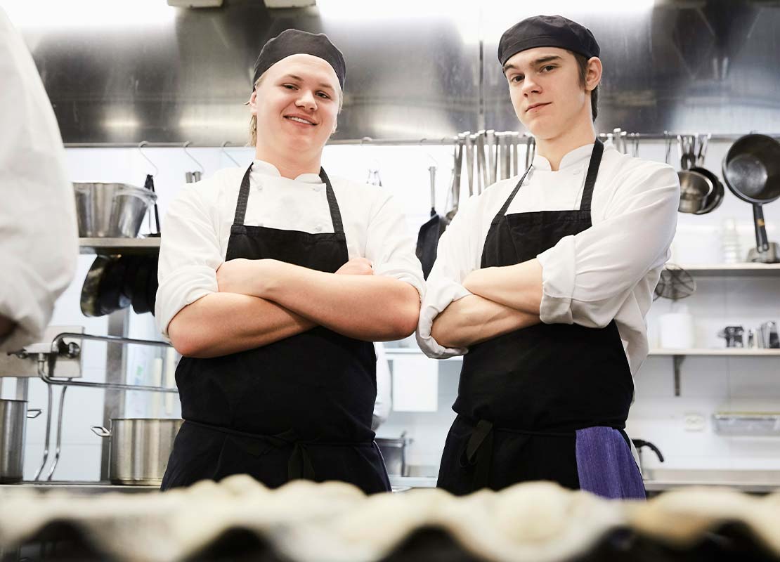 Dos chefs cruzados de brazos con gorro y delantal negro, al fondo utensilios de una cocina comercial
