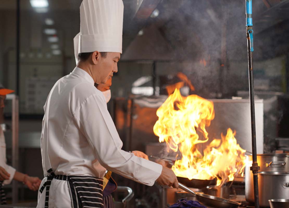 Chefs con gorra y delantal sostienen sartenes ardiendo sobre la estufa de una cocina comercial