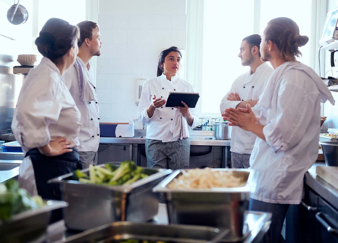 En una cocina comercial, chef sostiene una tablet y explica a 4 cocineros para capacitarlos