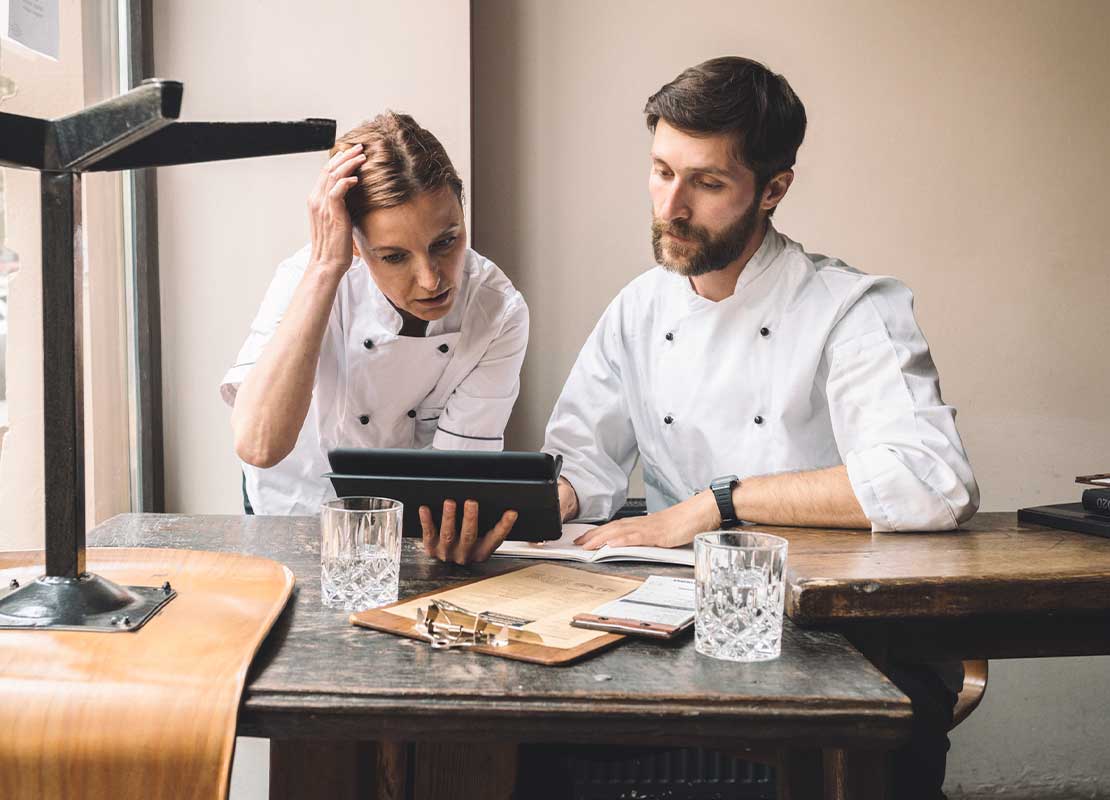 Dos chefs revisan una tablet, sentados a la mesa de una oficina sobre la que hay vasos