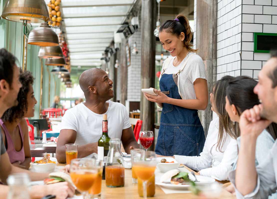 Mesera toma la orden de comensales sentados en una mesa con alimentos y bebidas servidos
