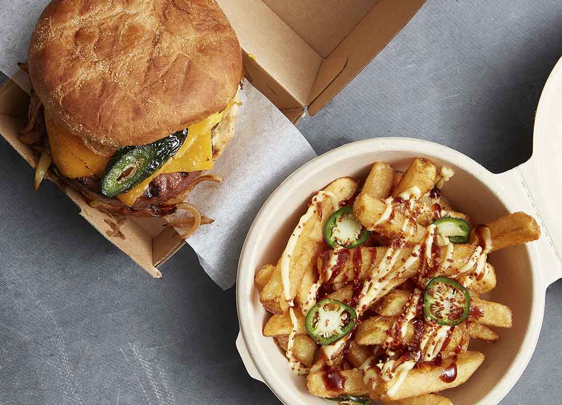 Una hamburguesa servida en una caja de comida y papas con salsa en un recipiente circular reciclable