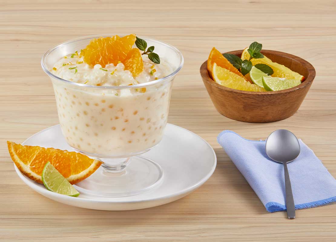 Copa de tapioca con leche condensada, coronada con naranja en gajos, al lado una taza con frutos cítricos