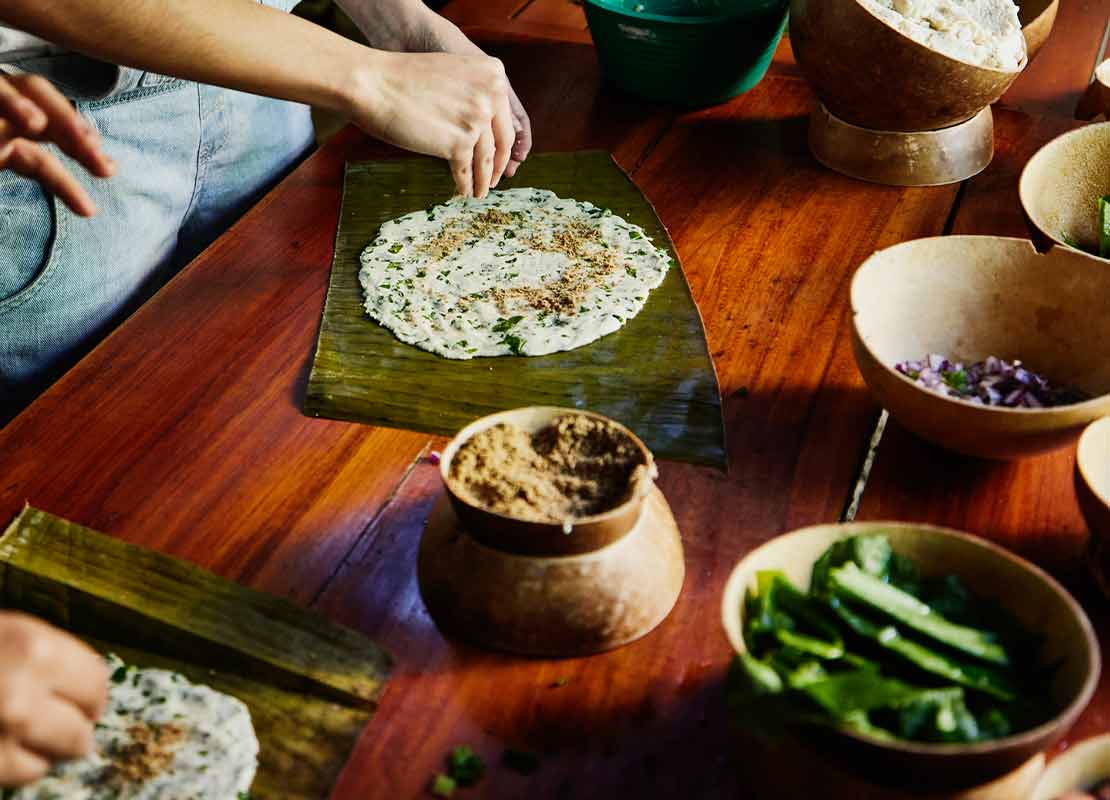Mesa con cuencos rodeada por personas que envuelven alimentos autóctonos en hojas de plátano