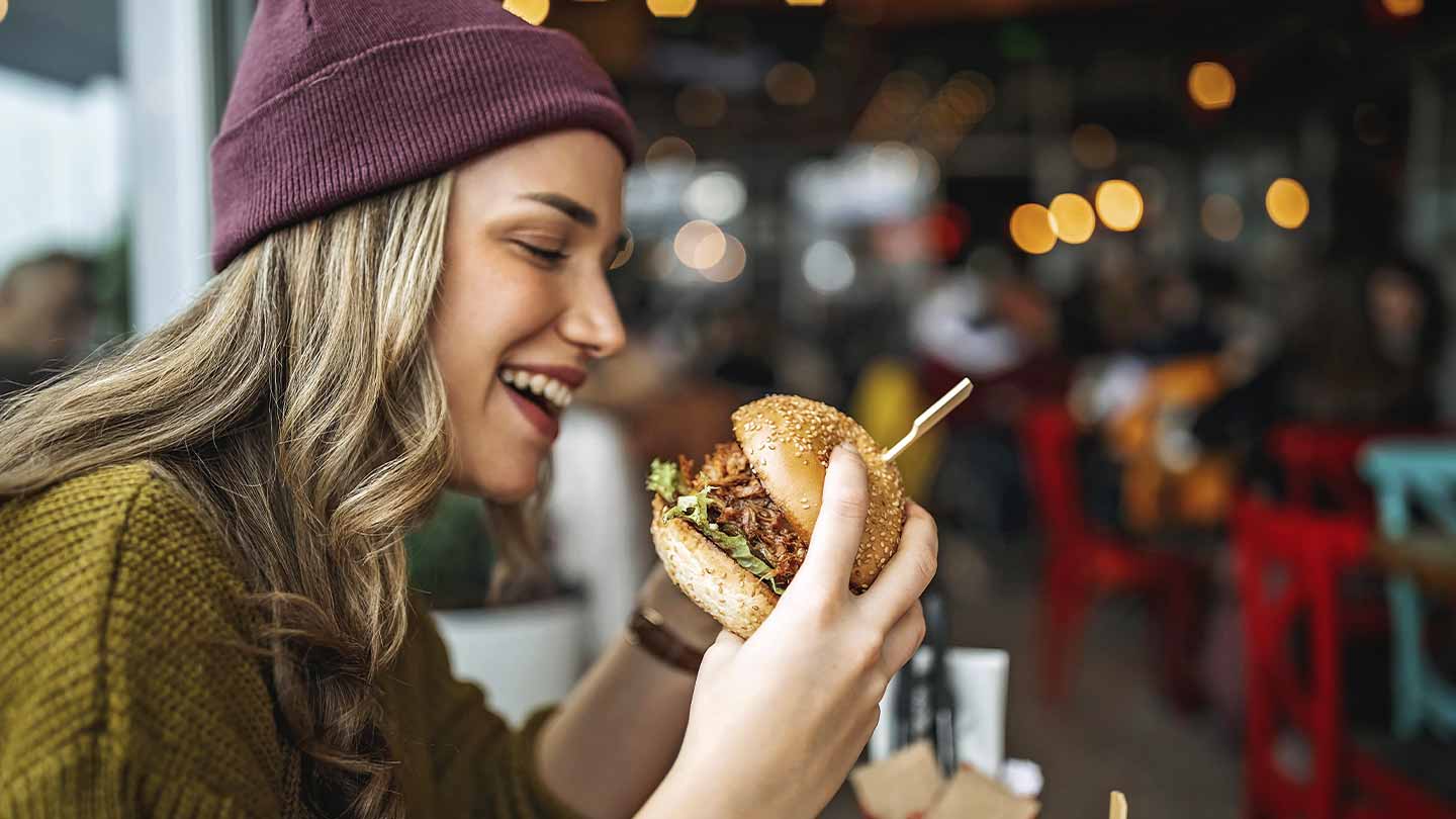 Mujer observa una hamburguesa que sostiene en sus manos, al lado bebida y otros apretivios