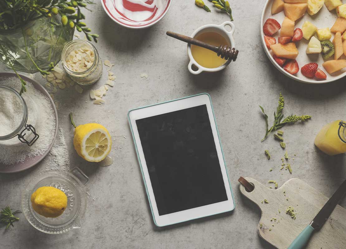 Tablet rodeada de recipientes con frutas, miel, avena, jugo, entre otros ingredientes sobre una mesa de cocina