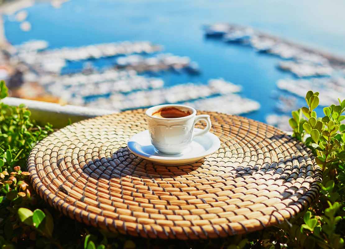 Taza de café servida sobre una mesa en un balcón, al fondo orilla del mar con casas y muelles