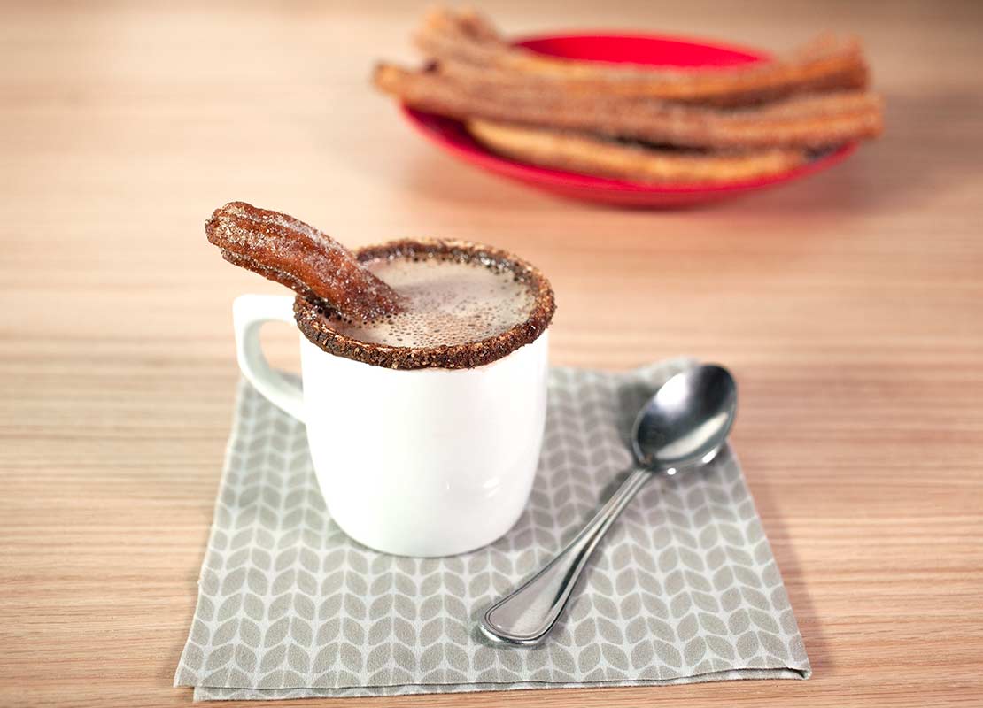 Taza blanca de chocolate caliente con el borde escarchado y un churro sumergido, al fondo, acompañamientos
