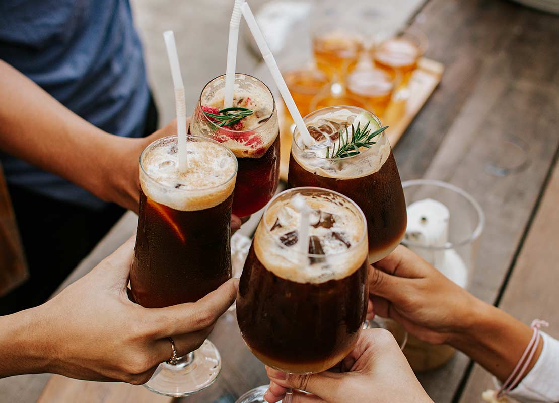 Manos sostienen 4 copas con bebidas craft, hielo y pajillas, las cuales chocan para brindar