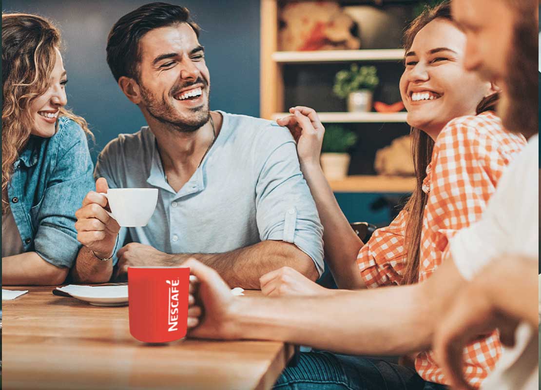 Grupo de 4 amigos sentados alrededor de una mesa sonríen y beben café en la taza de NESCAFÉ®