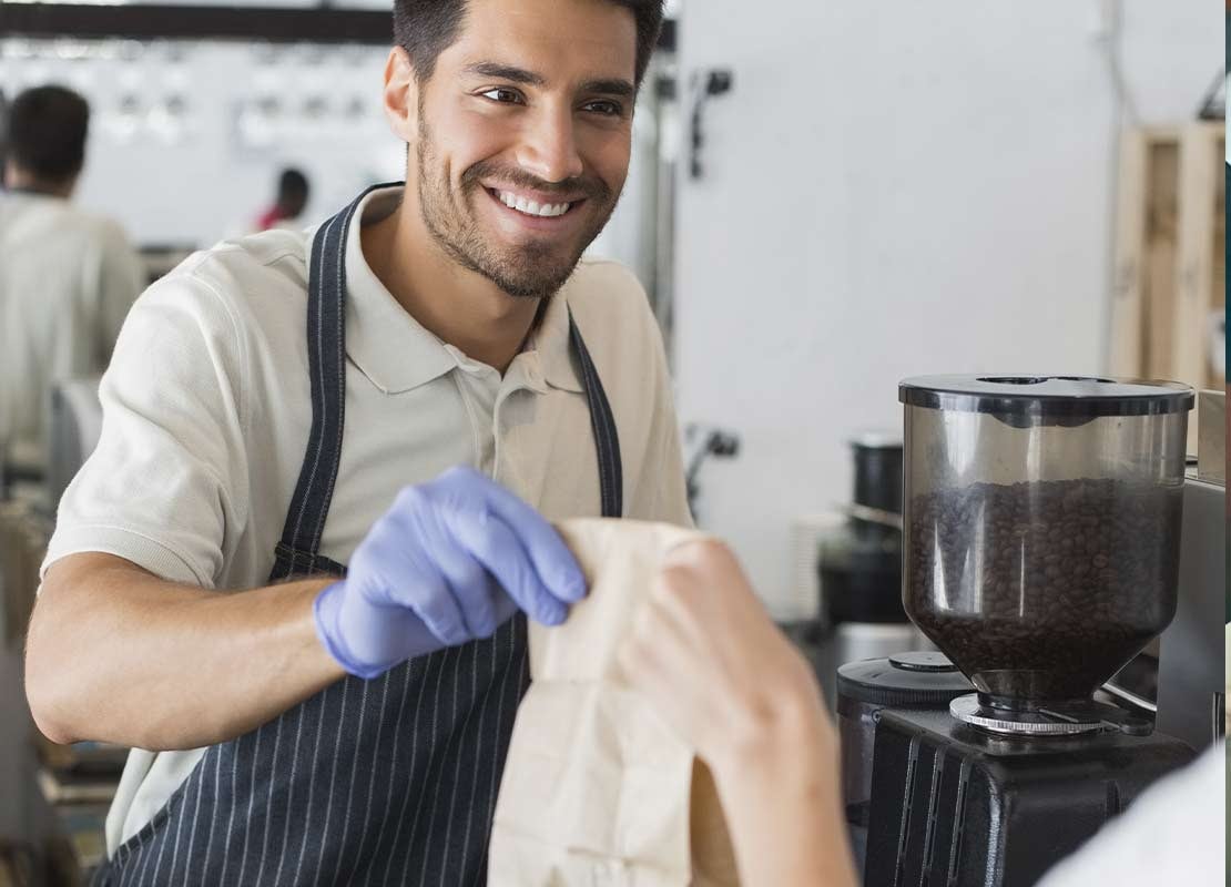 En una cafetería con máquinas de café al fondo, cajero entrega una bolsa de papel con comida a un cliente