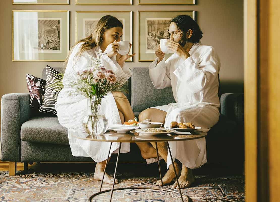 Dos huéspedes en bata sentadas en un sofá de una habitación de hotel disfrutan de un desayuno