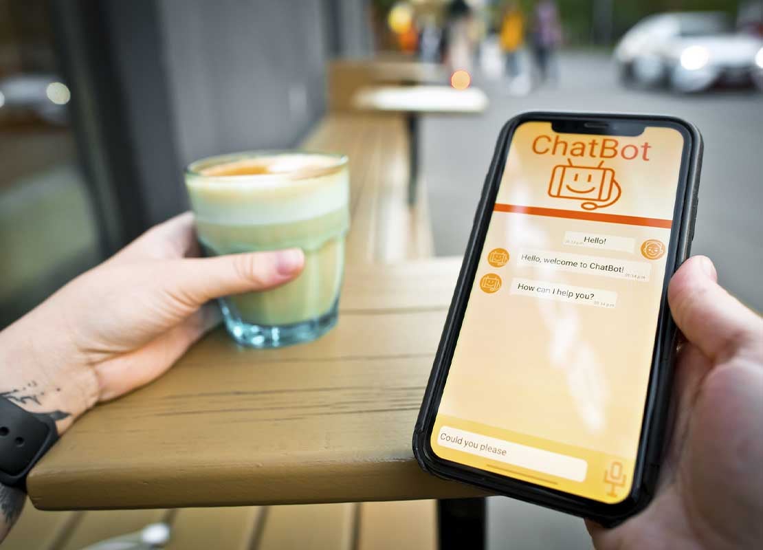 Mano sostiene un celular con un chatbot en su pantalla mientras con la otra sujeta un café