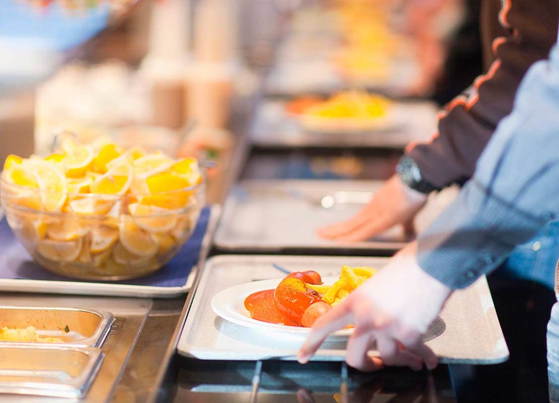 Clientes pasan bandeja con un plato de comida frente a una mesa de buffet con un tazón de naranja