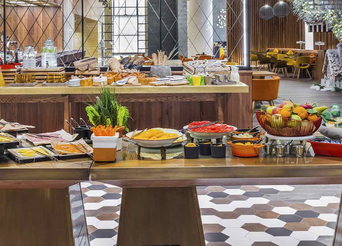 Salón con varias mesas de buffet con diversos alimentos como platos fuertes, ensaladas, bebidas, etc.